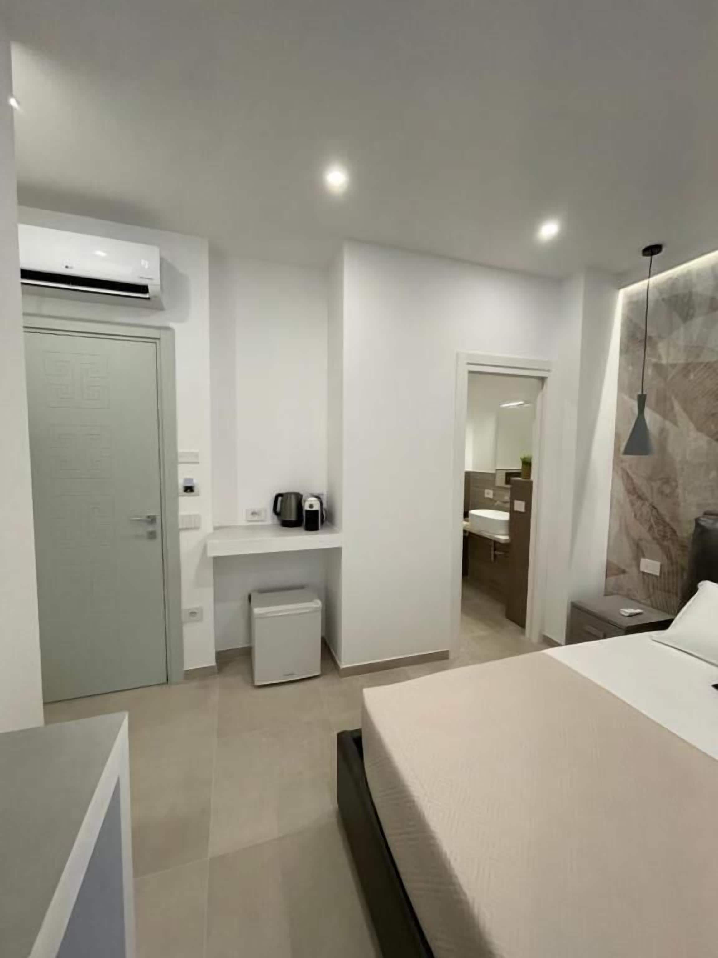 A modern hotel room with a bed, bedside table, and a small seating area. There is a bathroom visible through an open door.