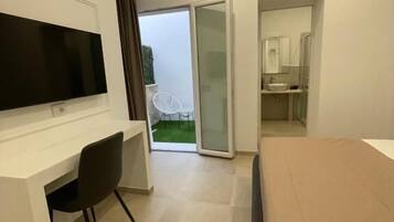 A modern hotel room with a flat-screen TV, a desk, a chair, a bed, a bathroom with a mirror, and a sliding glass door leading to an outdoor area.