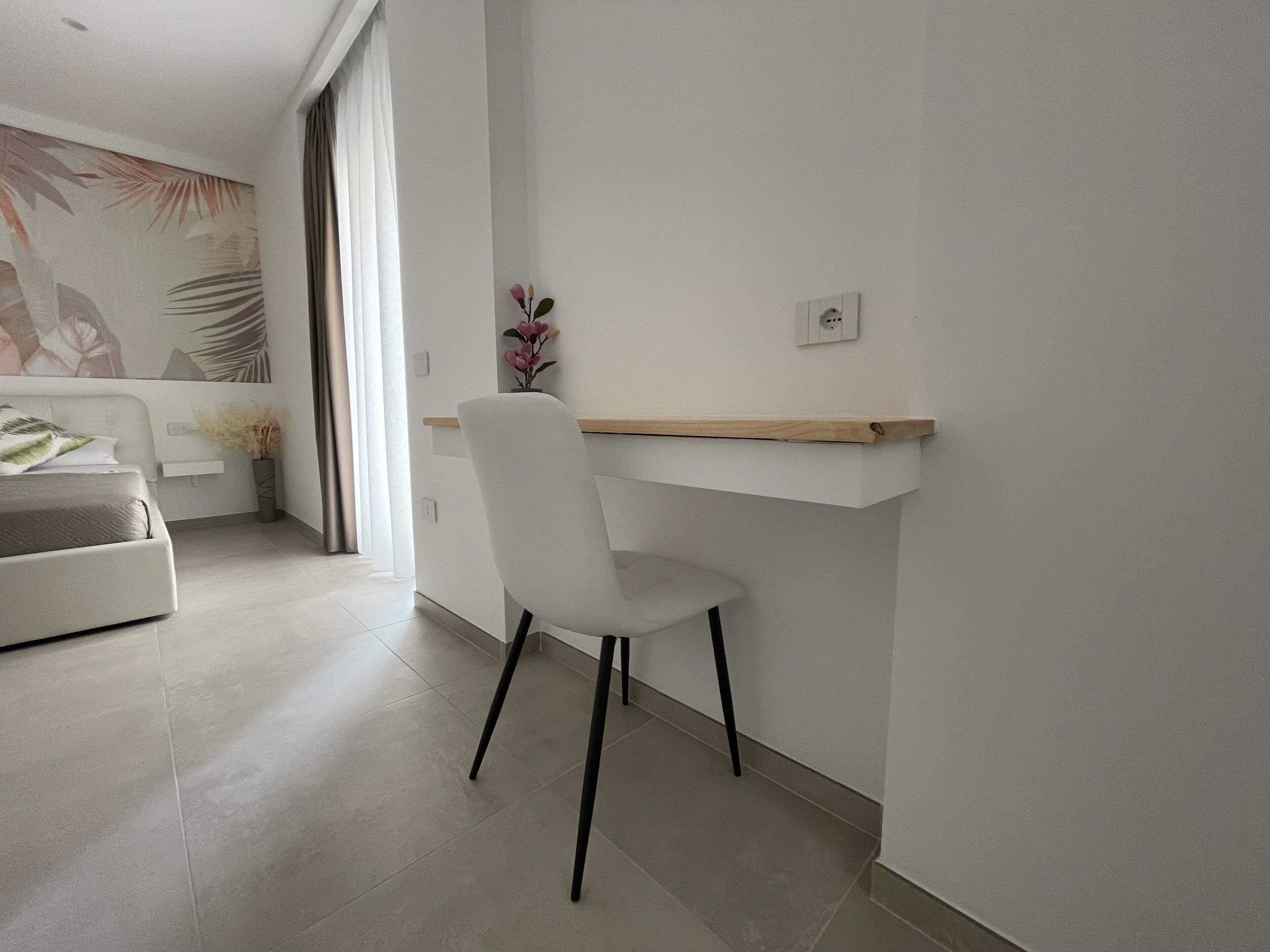 Ein moderner, minimalistischer Raum mit einem weißen Schreibtisch, einem weißen Stuhl und einem Bett mit grauem Kopfteil.