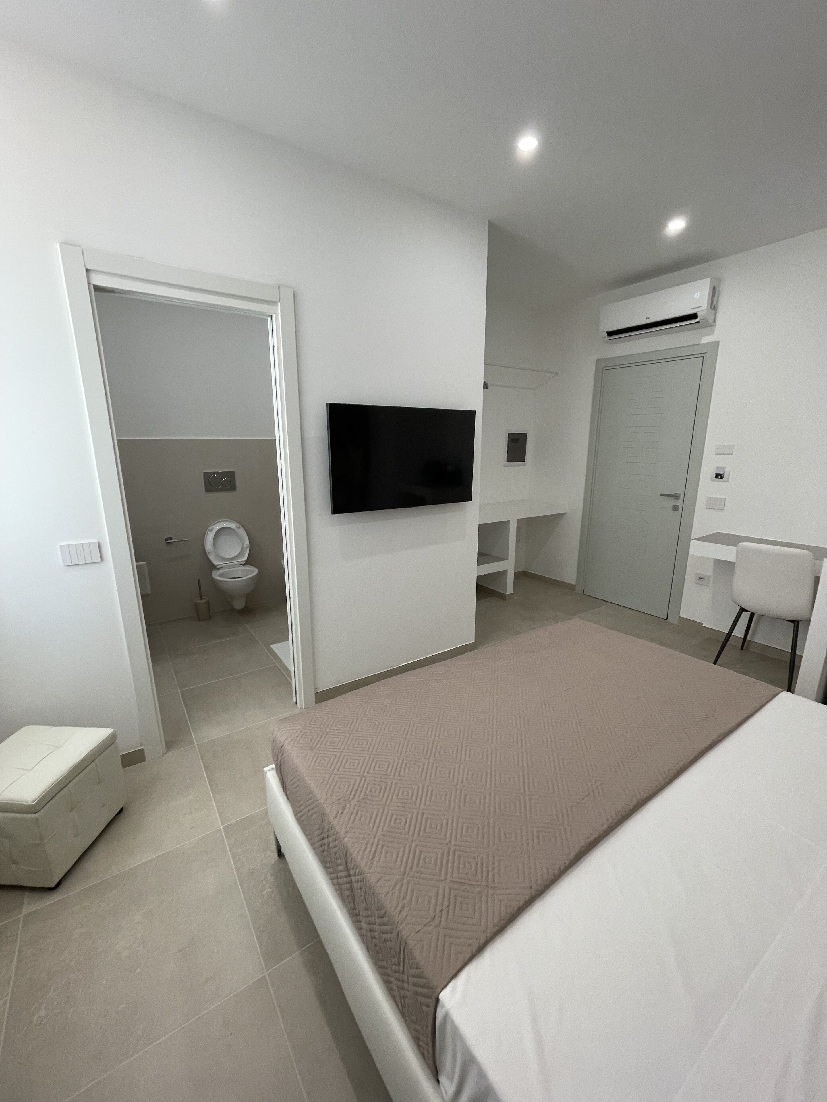 A modern hotel room with a large bed, a television mounted on the wall, a desk with a chair, and a separate bathroom with a toilet and sink.