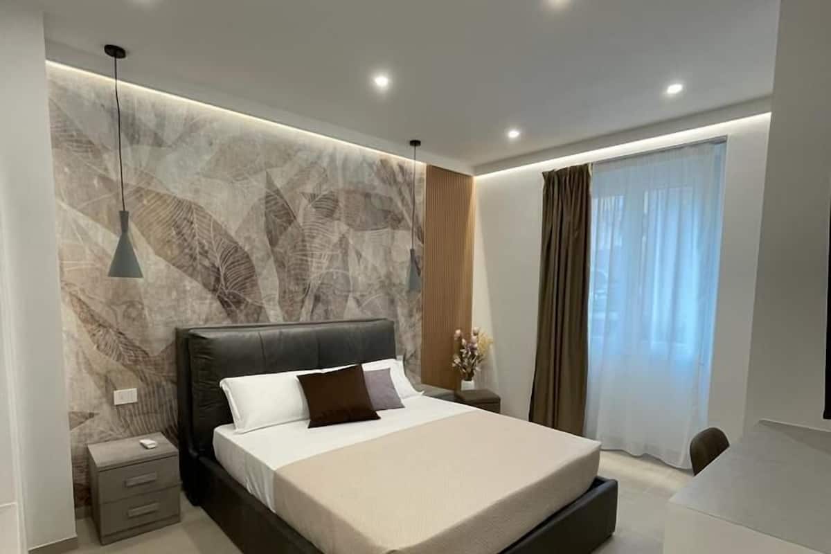 A modern bedroom with a large bed, a nightstand, a wall-mounted light, and a window with curtains.