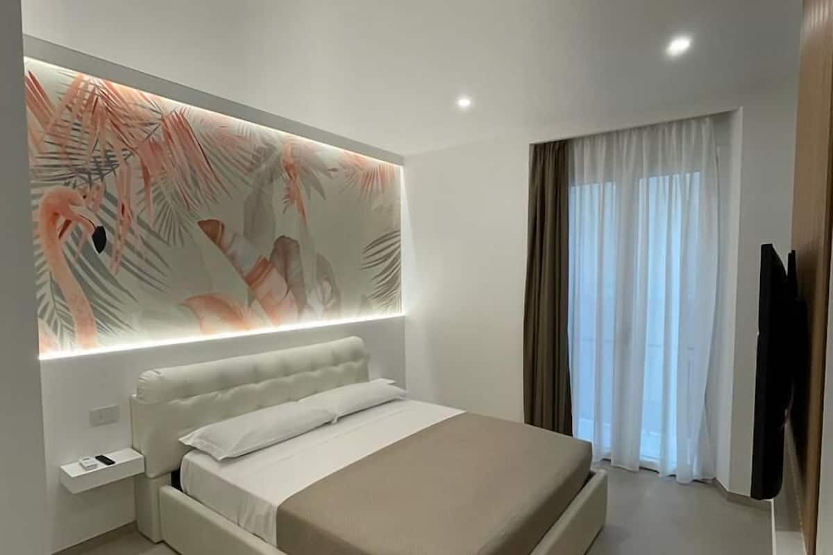 A modern bedroom with a large bed, a nightstand, a wall-mounted TV, and a window with curtains.