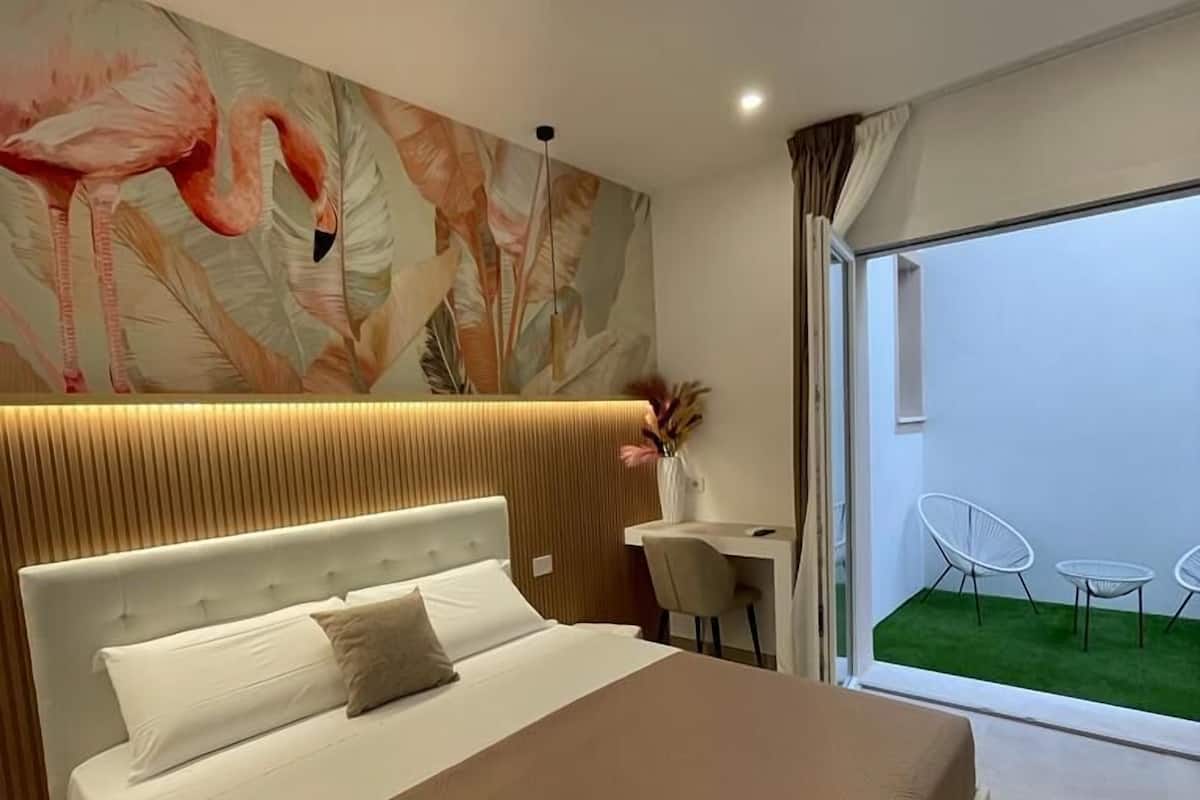 A modern bedroom with a large bed, a desk, and a view of a green lawn through a sliding door.