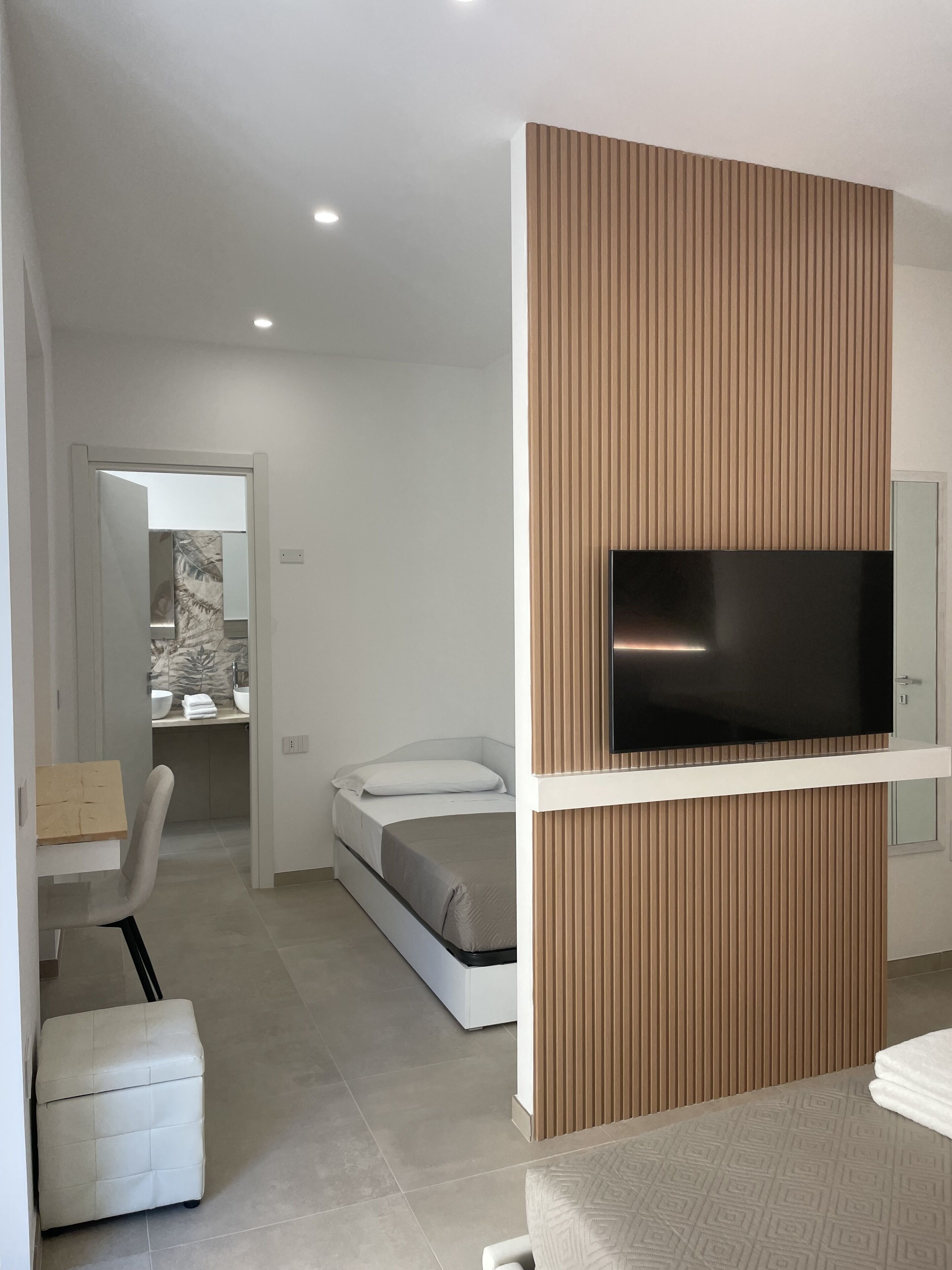 Un dormitorio moderno con cama, televisor montado en un panel de madera, un pequeño rincón de descanso y un baño visible a través de una puerta abierta.