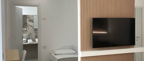 Un dormitorio moderno con cama, televisor montado en un panel de madera, un pequeño rincón de descanso y un baño visible a través de una puerta abierta.