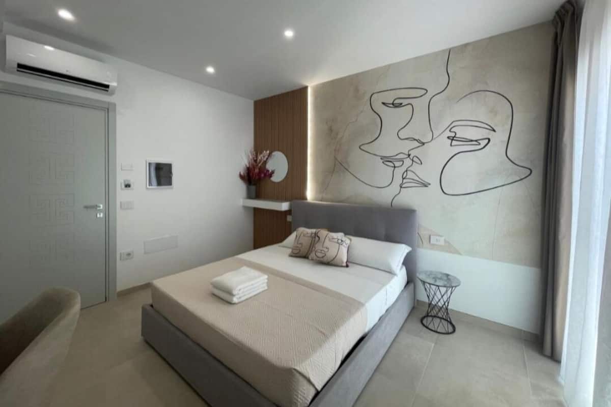A modern bedroom with a large bed, a wall-mounted air conditioner, a bedside table, and a decorative wall piece.