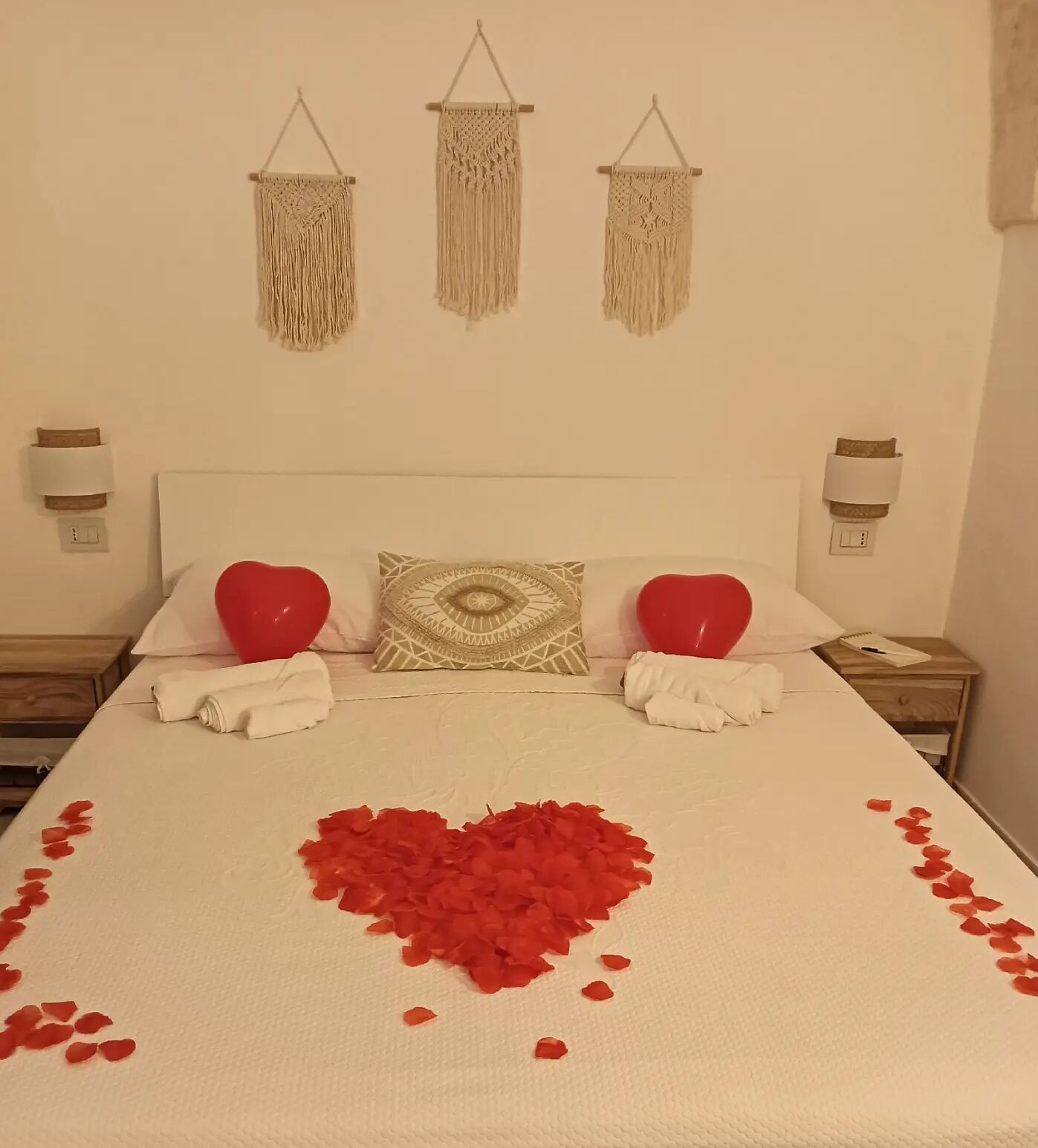 A neatly made bed with a heart-shaped arrangement of rose petals and two red heart-shaped pillows.