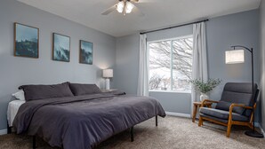 Une chambre à coucher comprenant un lit, une chaise, un ventilateur de plafond, une fenêtre avec des rideaux et des œuvres d’art accrochées au mur.