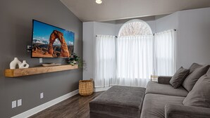 Un salon moderne avec un canapé gris, un téléviseur à écran plat fixé au mur, une étagère en bois ornée d’objets décoratifs et une grande fenêtre avec des rideaux légers.
