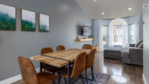 Un coin repas avec une table et des chaises en bois, un canapé gris et un téléviseur posé sur une étagère.