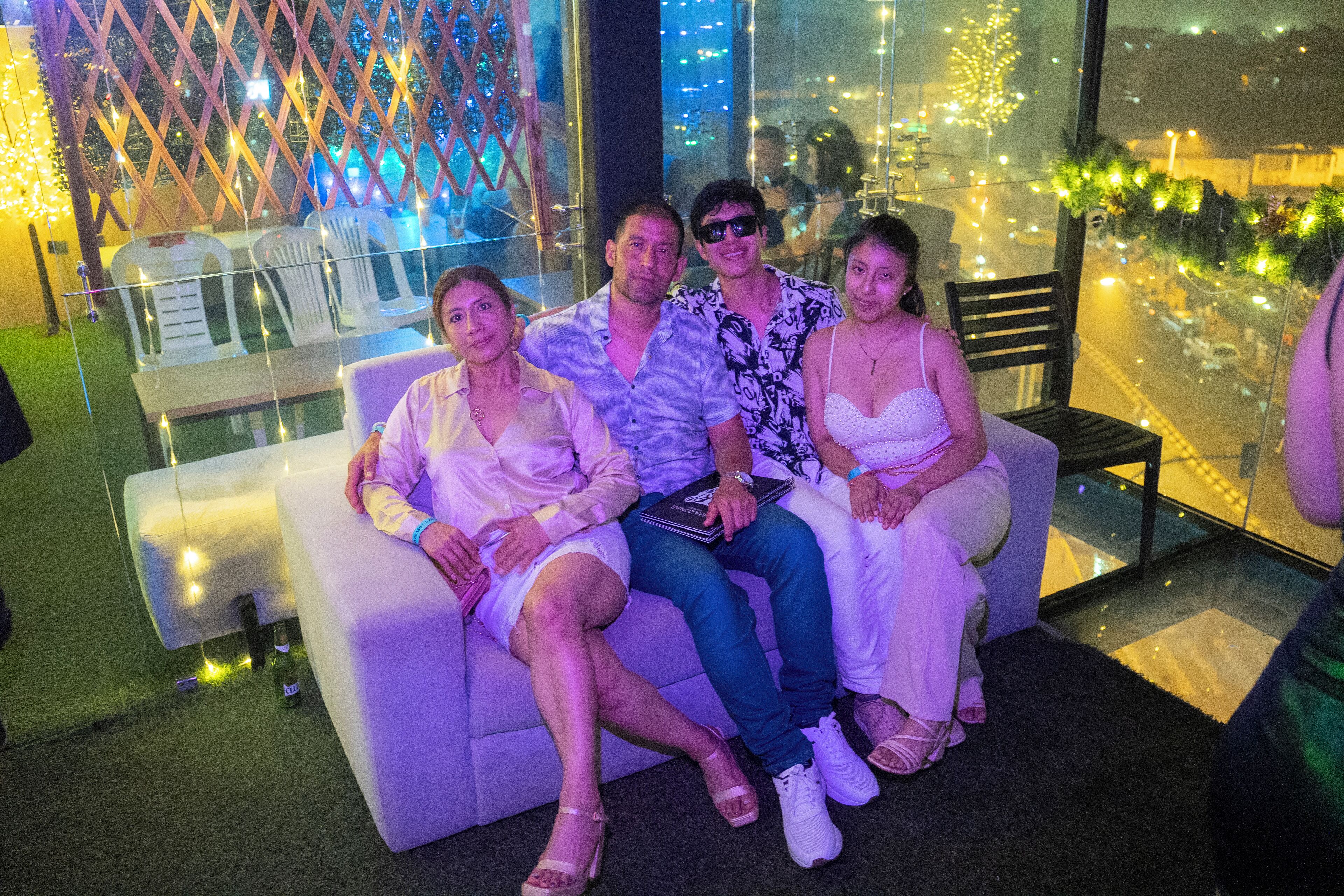 Un grupo de personas sentadas en un sofá blanco con un panel de vidrio detrás, con vistas a un paisaje urbano iluminado.