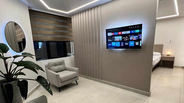 Una habitación de hotel moderna con un televisor de pantalla plana grande montado en la pared, un sofá cómodo, una planta en maceta y una mesita de noche con una lámpara.