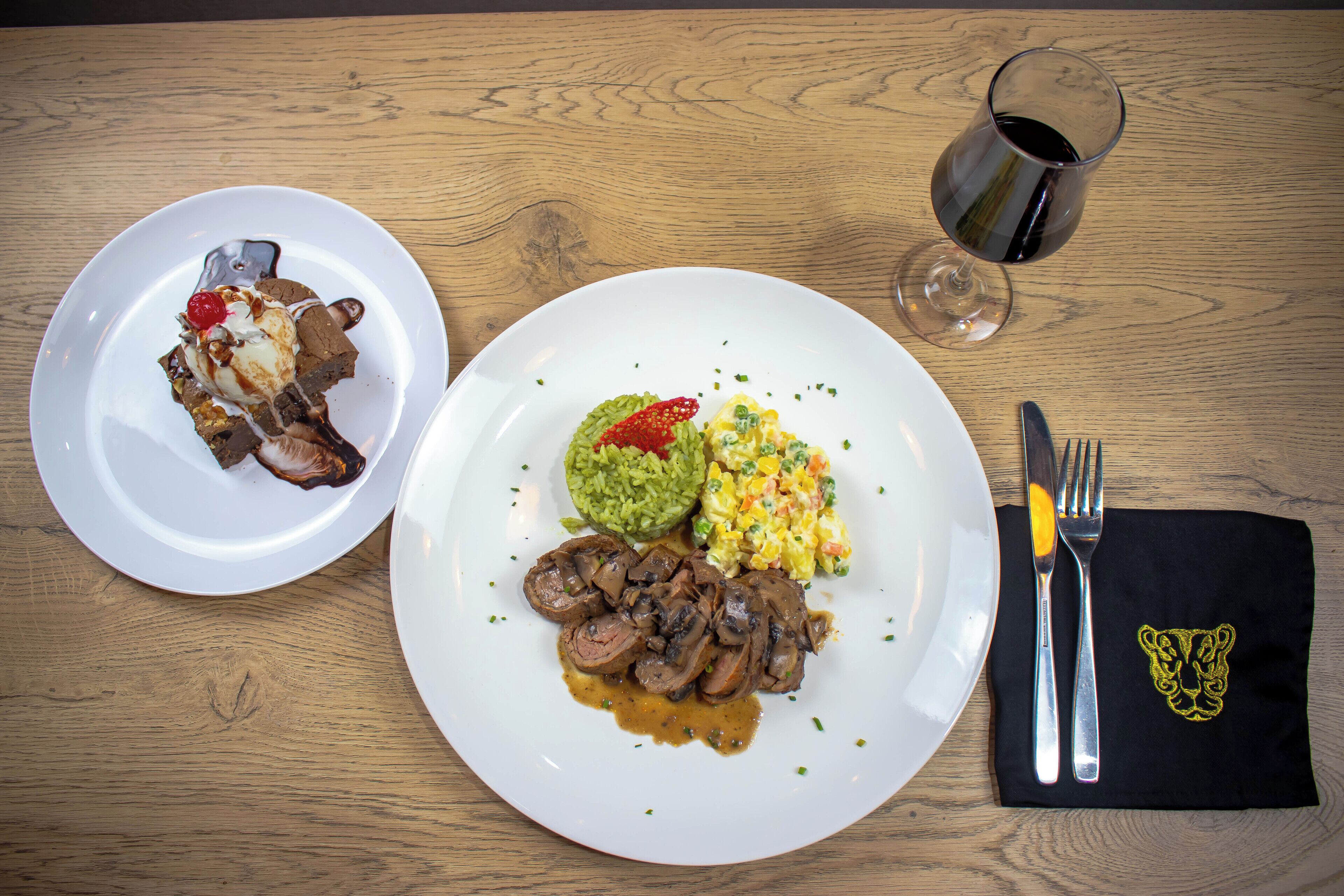 Two plates of food on a wooden table with a glass of red wine and cutlery.