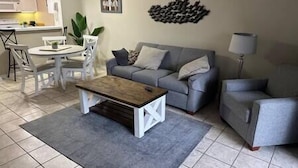 A living room with a grey sofa, a wooden coffee table, and a grey armchair. There is a dining area with a white table and chairs. - Beautiful Condo in Ocean Creek (Horry County)