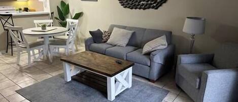 Un salon avec un canapé gris, une table basse en bois et un fauteuil gris. On y trouve également un coin repas avec une table et des chaises blanches.