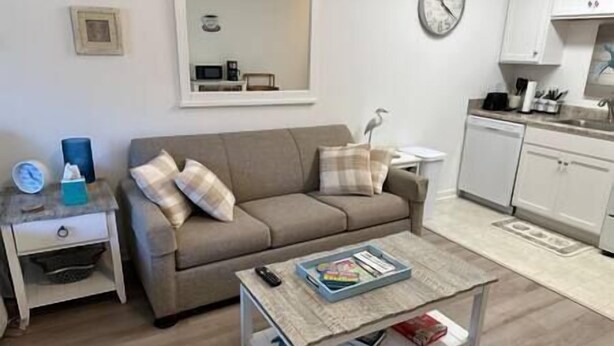 Una sala de estar con un sofá, una mesa de centro con libros, una mesita auxiliar y una zona de cocina con armarios y un reloj.