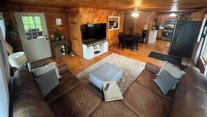 A cozy cabin interior with a wooden ceiling, a TV in the living room, a dining area, and a kitchen with modern appliances.