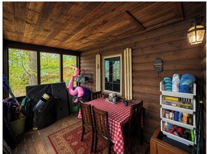 A wooden cabin with a checkered table, chairs, a red rug, a shelf with items, and a window with a view of trees.