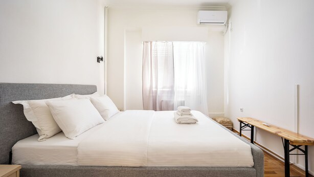 Una cama bien tendida con sábanas blancas, un cabecero gris y un banco de madera a su lado.