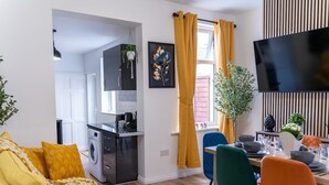 A modern living room with a sofa, a dining area with chairs and a table, a kitchen with appliances, and a TV mounted on the wall. - Open Mind Property - Luxury 5 Beds & Free Parking (Portsmouth)