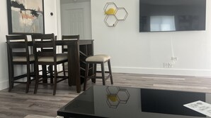 A modern living room with a dining area, featuring a black coffee table, dark wooden chairs, a TV mounted on the wall, and hexagonal wall decor. - Boca Jewels with Heated Jacuzzi (Boca Raton)