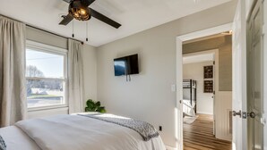 Une chambre avec un grand lit, un ventilateur de plafond, un téléviseur fixé au mur et une fenêtre avec des rideaux.