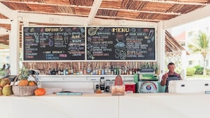 A bar area with a chalkboard menu and drinks list, a cash register, and a tropical fruit display. - Magic Cortesea luxury beach (Punta Cana)