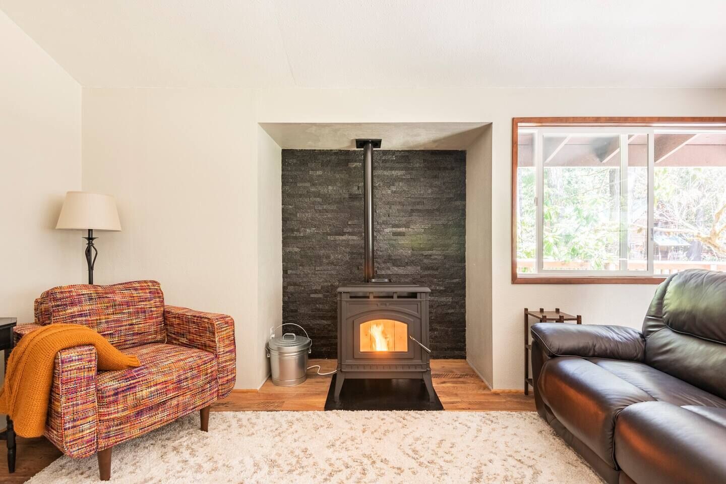 Una sala de estar acogedora con una chimenea de piedra, un sillón colorido, un sofá de cuero negro y un ventanal grande.