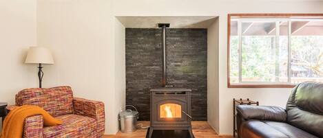 Una sala de estar acogedora con una chimenea de piedra, un sillón colorido, un sofá de cuero negro y un ventanal grande.