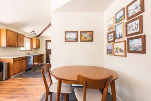 A modern kitchen with wooden cabinets, a built-in dishwasher, and a dining area with a round wooden table and two chairs. - Lupine Glen: Community Pool, Wifi, Fire, Deck! (Rhododendron)