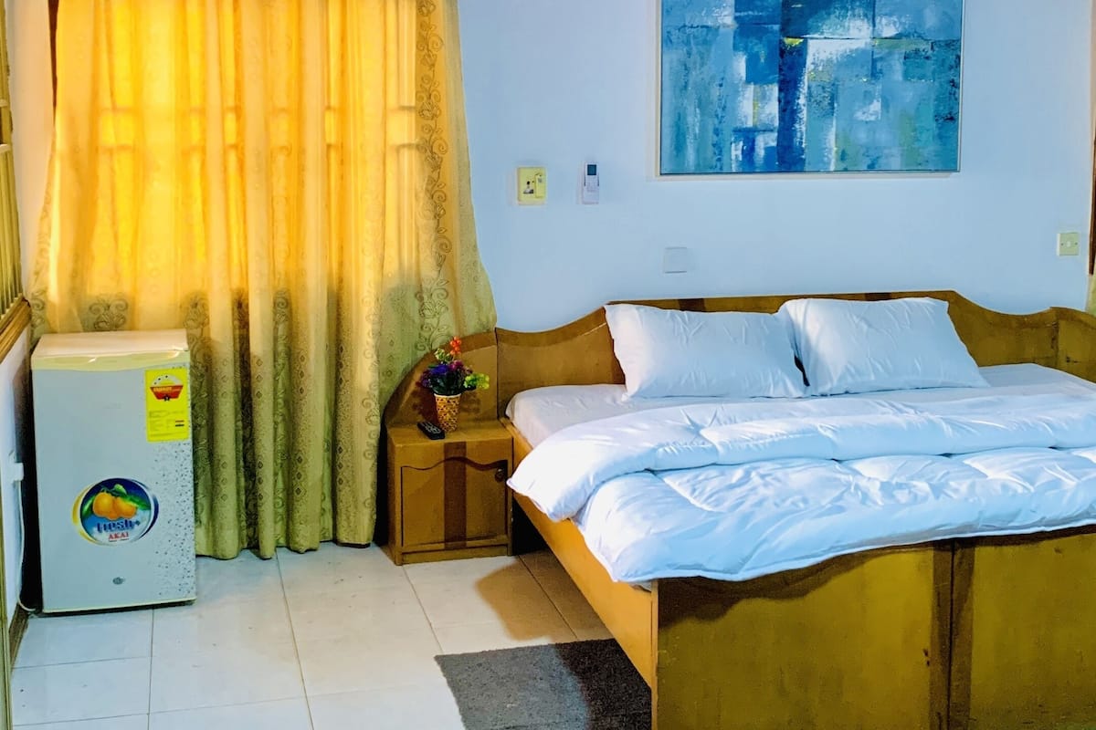 A bedroom with a bed, a nightstand, a wall-mounted lamp, a refrigerator, and a painting on the wall.