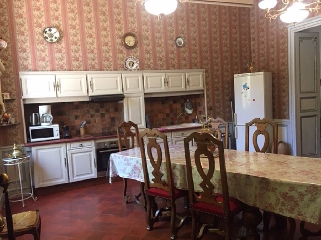 Une cuisine avec des meubles blancs, un four à micro-ondes et un réfrigérateur. On y trouve une table à manger avec des chaises et une nappe à motifs.