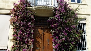 Un edificio con puerta de madera, balcón con barandilla de hierro forjado y enredaderas florecientes de color morado.