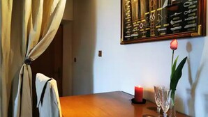 Une table en bois avec une bougie rouge et deux verres transparents, un menu encadré accroché au mur et un rideau noué dans un coin.