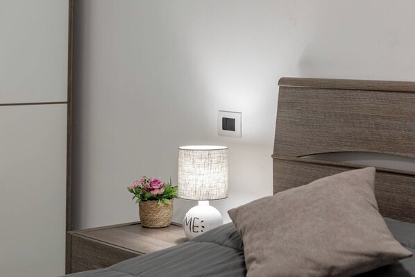Un lit bien fait, avec un oreiller gris, une lampe de chevet et un petit panier tressé contenant des fleurs roses.