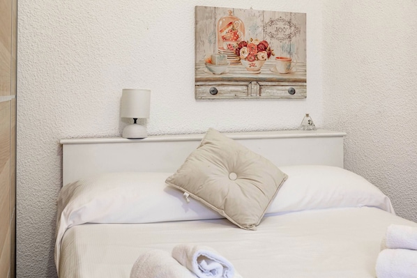 Een net opgemaakt bed met één kussen en een opgevouwen handdoek, een wandlamp en een decoratief schilderij boven het bed.