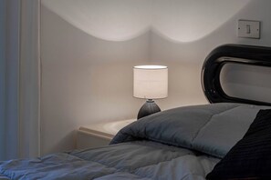 Een bed met een grijs en zwart dekbed, een bedlamp met een witte lampenkap en een stopcontact.
