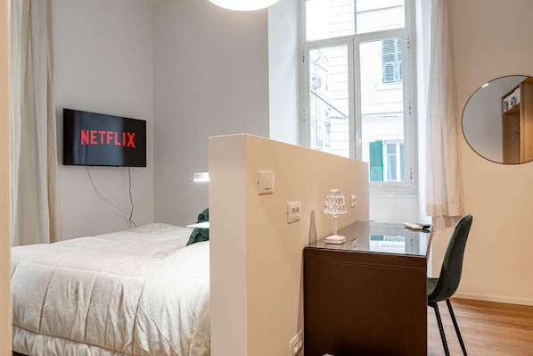 A hotel room with a bed, a desk, a chair, a TV mounted on the wall, and a window. - THE QUEEN'S APARTMENT - Ariston - Centro (Sanremo)