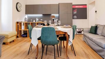 Un espace repas moderne avec une table en bois dressée pour quatre personnes, entourée de chaises de couleur turquoise. On aperçoit à l'arrière-plan une cuisine équipée d'un four encastré et d'une horloge murale.