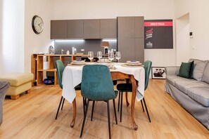 Un espace repas moderne avec une table en bois dressée pour quatre personnes, entourée de chaises de couleur turquoise. On aperçoit à l'arrière-plan une cuisine équipée d'un four encastré et d'une horloge murale.