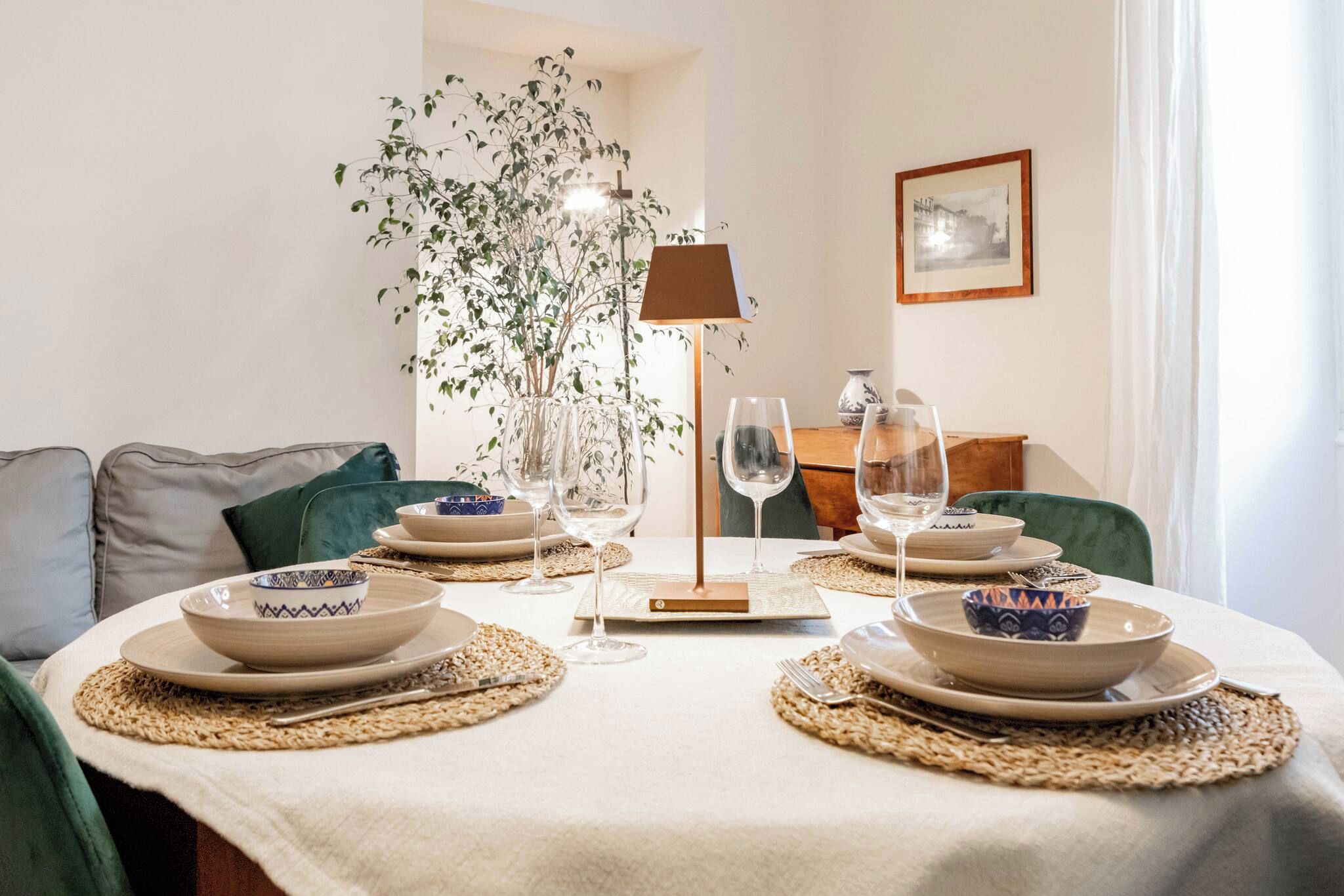A dining table set with plates, bowls, and wine glasses, surrounded by chairs and a plant.