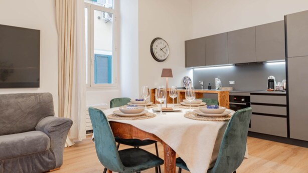 A modern living room with a grey sofa, a dining table set with plates, glasses, and a clock on the wall.