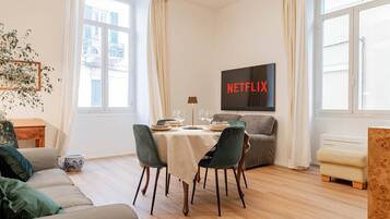A modern living room with a dining area, a sofa, a TV mounted on the wall, and a window with curtains.