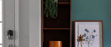 Une console en bois, ornée d’un vase et d’un tableau encadré, est placée contre un mur de couleur turquoise. Un bol se trouve sur l’étagère inférieure, et une porte, dotée d’une poignée et d’une serrure, complète l’ensemble.