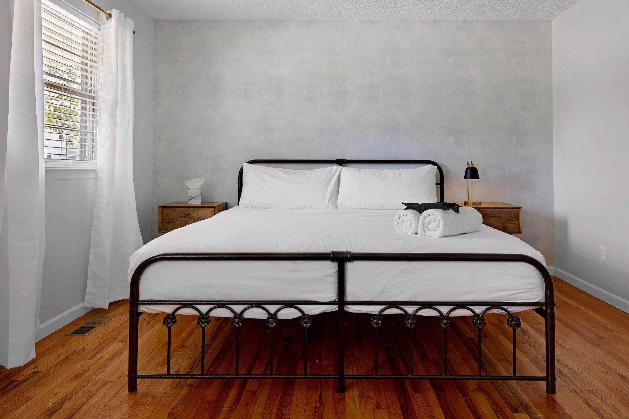 A neatly made bed with white linens and a black metal frame, a bedside table with a lamp, and a window with white curtains.