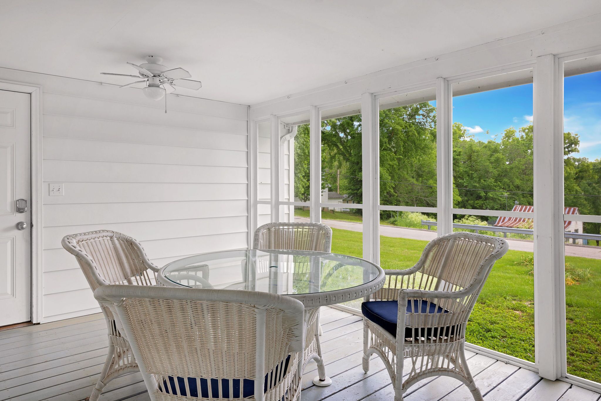 A covered porch with a glass table and wicker chairs, overlooking a green landscape.