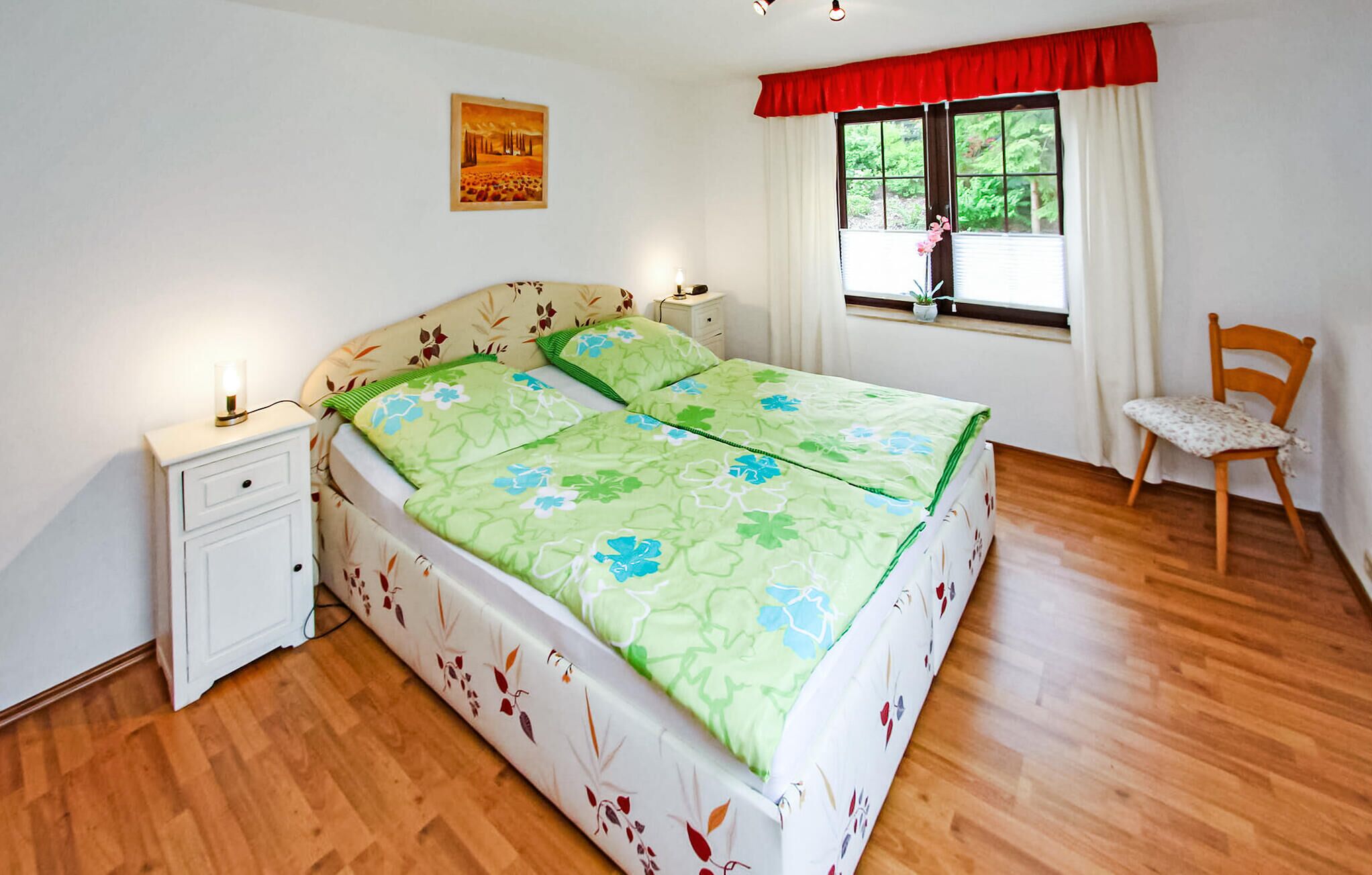 A bedroom with a bed, nightstands, a chair, a window with curtains, and a painting on the wall.