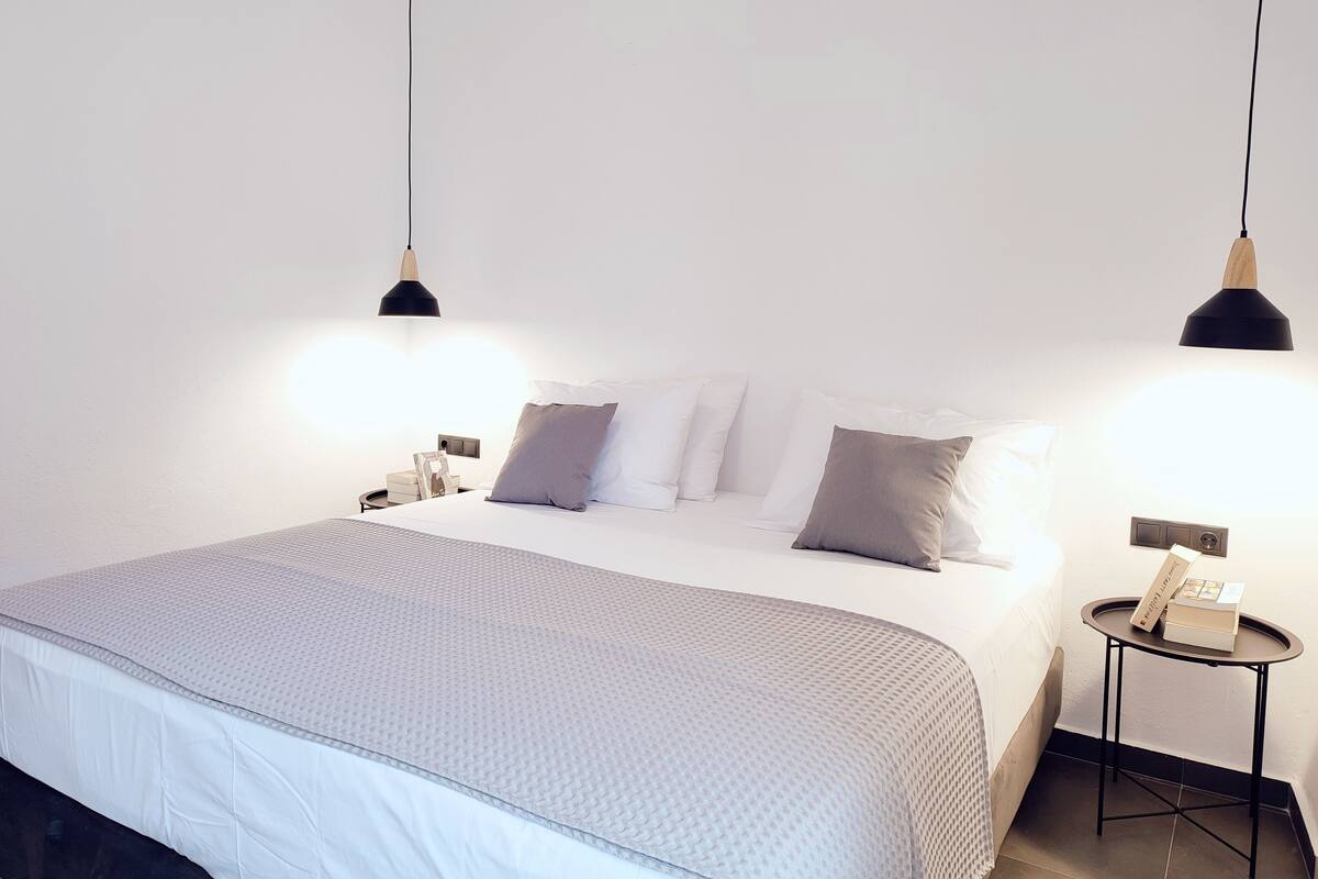Een netjes opgemaakt bed met een grijs-witte dekbedovertrek, twee nachtkastjes met lampen, en een klein rond tafeltje met een boek aan de zijkant.