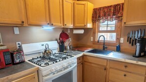 A kitchen with wooden cabinets, a gas stove, a sink, and a window with a floral valance. - Bluff Bungalow w/ Patio, Dogs Welcome (Bluff)