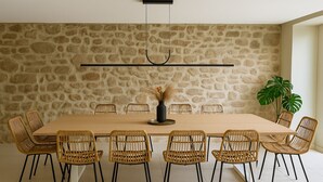 A dining area with a stone wall, a long wooden table, wicker chairs, and a hanging light fixture. - Gîte 12 Personnes, Sauna, Billard, Baby-foot, Bain à Remous, Terrasse, Barbecue (Lapoutroie)